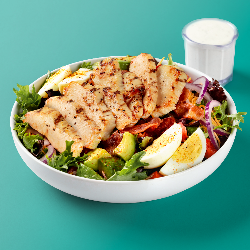 Cobb salad with bacon, boiled egg, avocado, cheese, onions, and chicken on a bed of crunchy lettuce with tasty ranch dressing on the side.